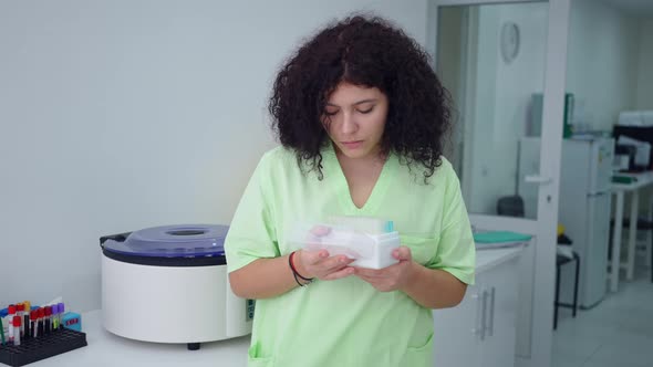 Concentrated Plussize Lab Assistant Examining Test Tubes Box and Looking at Camera Smiling