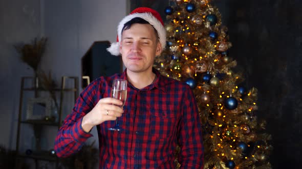 Young Man in Santa Hat Drinks Champagne Standing Near Christmas Tree in Dark Room