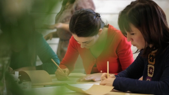 Three Women Sit Around Wooden Table And Draw Sketches In Art Studio