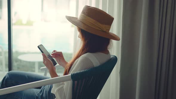 Happy young woman sit on a armchair near the window at hotel while use mobile phone communication