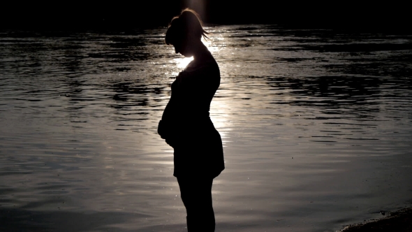 Pregnant Woman Pats Her Belly On Sunset On River Background