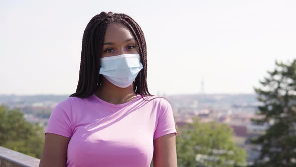 A Young Black Woman in a Face Mask Looks at the Camera - a Cityscape in the Blurry Background
