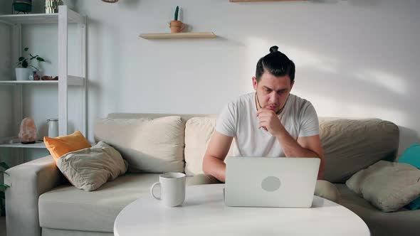 Young Unhappy Man Using Laptop Typing and Working Sitting on the Couch at Home