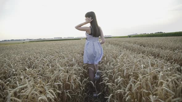 Young Lady in Dress Walks Among Golden Wheat Field and Poses with Hat