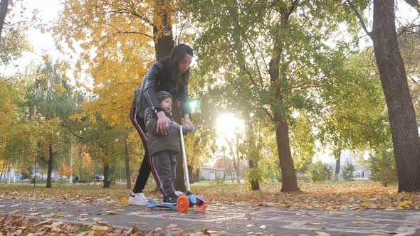 Young Mother Showing Her Toddler Son How To Ride a Scooter in a Autumn Park. Active Family Leisure