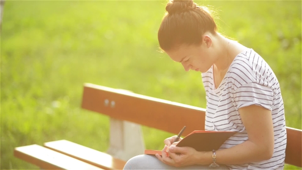 Beautiful Young Woman Is Writing a Diary Outdoors In The Park, Student Studying On a Bench