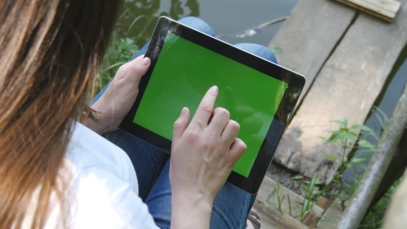 Female Hands Using Digital Tablet Pc With Green Screen Sitting On a Wooden Jetty By The Lake.