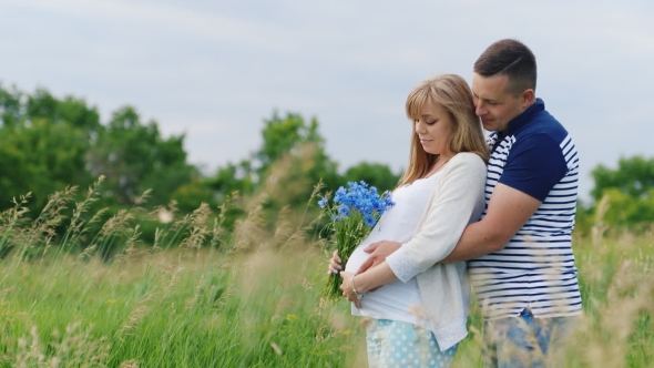 Happy Family - Man Hugging His Pregnant Wife From Behind. It Is In The Park Or Field