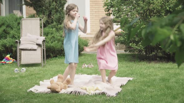 Two Cheerful Little Girls Taking Each Other's Hands and Start Jumping on Blanket Outdoors. Wide Shot