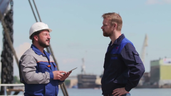 Two Skilled Workers At a Port With a Tablet PC, Nearby Cargo Ship Partly Visible