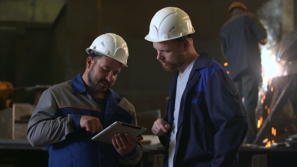 Two Engineers In Hardhats Discussing In Front Of Welding Process