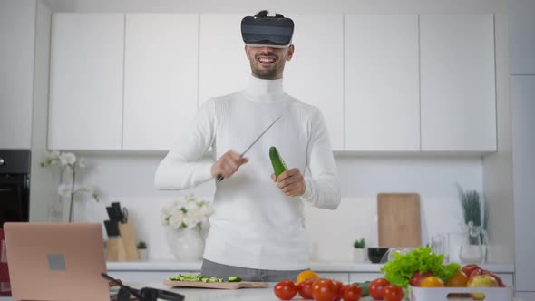 Laughing Funny Middle Eastern Young Man in VR Headset Cutting Cucumber with Knife in Kitchen