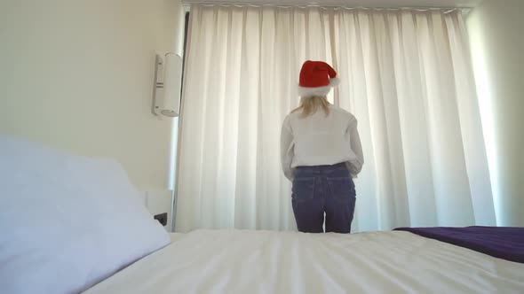 a woman in a Santa Claus hat gets out of bed and opens the window curtains.