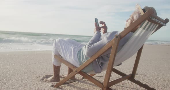 Hispanic senior woman relaxing on sunbed on beach at sunset, using smartphone