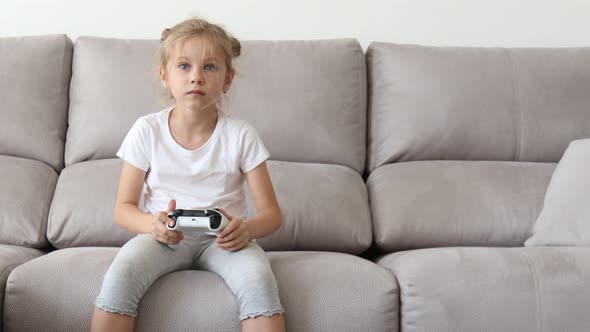 Enthusiastic Kid in Casual Outfit Playing with Joystick While Sitting on Sofa in Livingroom