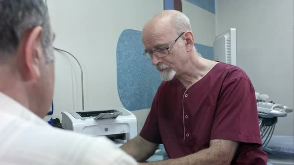 Mature Doctor at His Desk, Make Some Notes, Listening To a Senior Patient