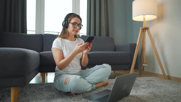 Casually Dressed Woman with Headphones is Sitting on Carpet with Smartphone and Laptop and Working