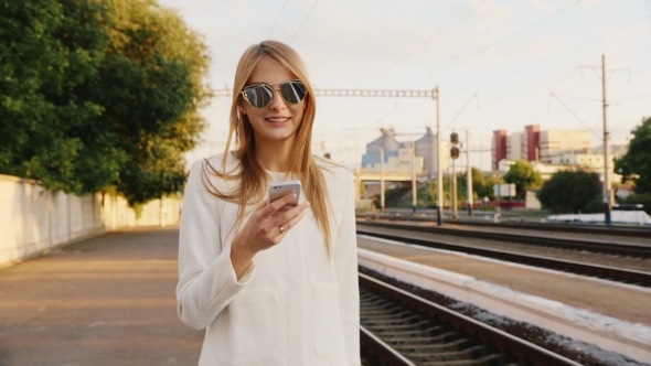 Young Woman In Sunglasses Smiling, Talking On The Peron Along The Railway. Use Smartphones