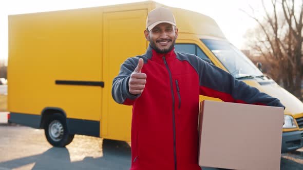 Smiling Adult Delivery Man Carrying Cardboard Box Showing Thumb Up Gesture and Looking at Camera