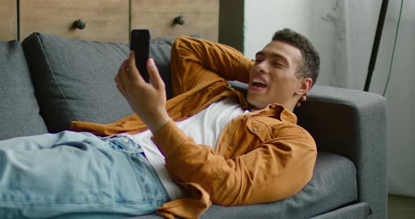 Hispanic Man Is Lying on Sofa, Having Video Chat on Smartphone