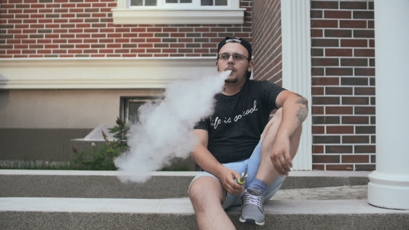Young Man Sitting Near House On The Street And Smoke E-Cigarette. Vape. 