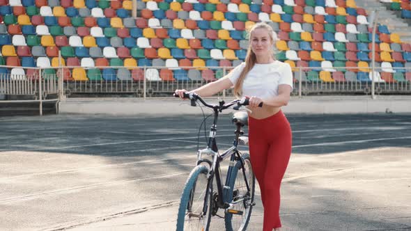 Athletic Woman in Red Leggings and White T-shirt Leading a Bicycle on a Stadium Track