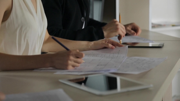 Female Hands Holding a Plan Of Building, Sitting Next To Man And Designs On Paper.