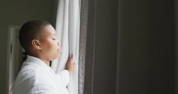 Happy african american plus size woman wearing robe, opening curtains and looking through window