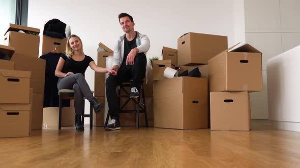 A Happy Moving Couple Sits on Chairs in an Empty Apartment, Holds Hands and Smiles at the Camera