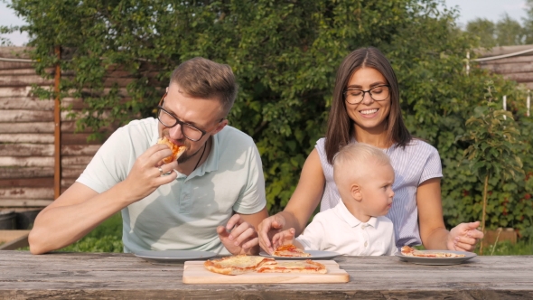 Young Happy Family Having Picnic In The Garden. Mom, Dad And Son Eating Pizza Outdoors