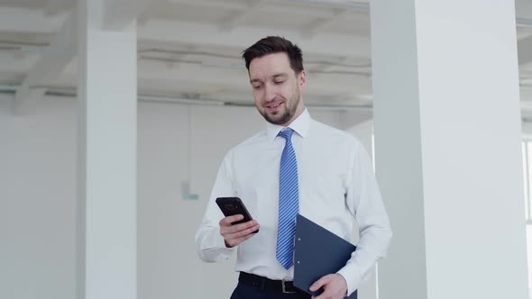 Businessman Man Stands with Tablet in Hand and Looks Into Smartphone and Smiles Front View