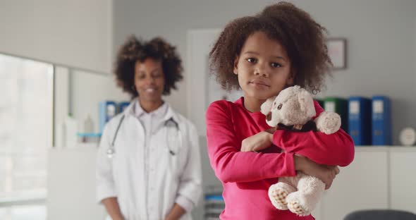 Portrait of Little African Girl Holding Teddy Bear with Young Female Doctor Standing on Background