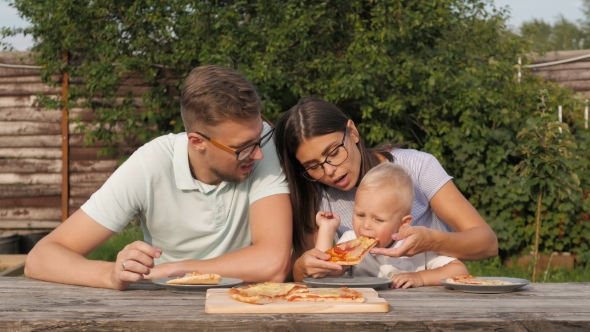 Young Happy Family Having Picnic In The Garden. Mom, Dad And Son Eating Pizza Outdoors