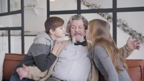 Grandchildren Coming To Grandfather Sitting on Couch and Kissing Senior Man on Cheeks, Portrait