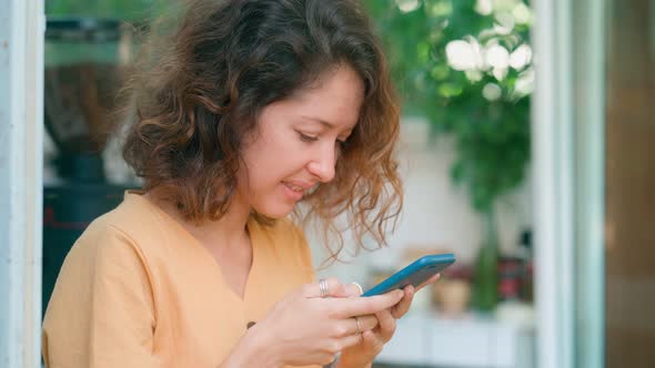 Smiling Curly Woman in Yellow Dress Using Her Phone Happily While Sits in Coffee Shop