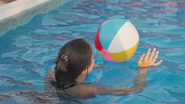 A Girl in a Bright Bathing Suit Swims with an Inflatable Ball in a Pool with Clear Water on a Summer