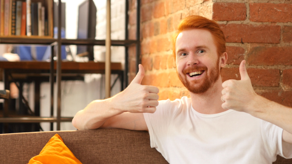 Both Hands Thumbs Up by Smiling Man w/ Red Hairs and Beard, Sitting on Sofa 