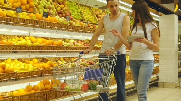 Young Caucasian Couple Walking In a Supermarket With a Market Trolley And Choosing Fresh Apples. Man