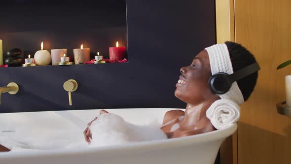 Smiling african american woman with towel taking bath and wearing headphones in bathroom