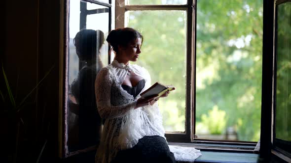 Beautiful Elegant Woman is Reading Book Sitting on Windowsill of Old Village House in Summer