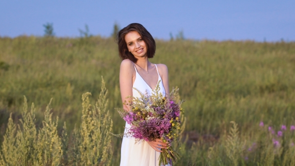 Young Woman Standing On Green Field With Flower Bunch And Smiling