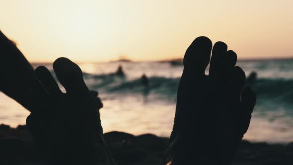 POV Silhouette of Feet of Young Man Lying on Sandy Beach By Ocean During Sunset
