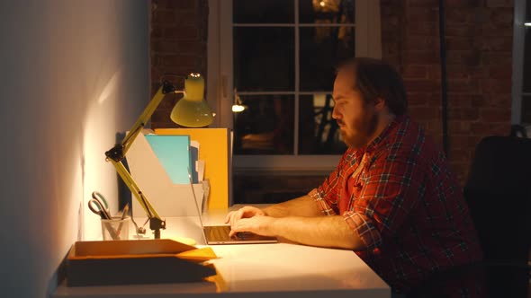 Young Man Sitting at Table and Working on Computer Late in Evening While Neighbor Sleeping at Desk