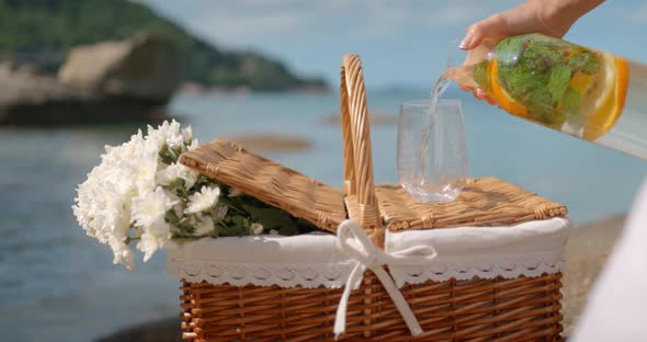 Lemonade with Mint Pouring Into Glass Standing on Picnic Basket with Flowers on Seashore Outdoors