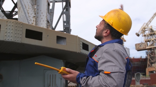 Dock Worker With Yellow Helmet And Walkie-talkie Directs The Cargo Crane In Shipping Port