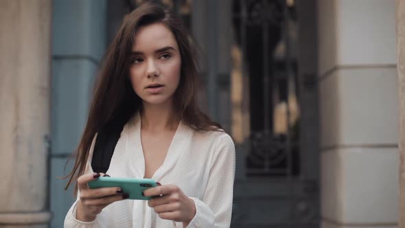 Pretty Young Student Brunette Girl with a Bag on Her Shoulder Holding and Using Her Smartphone in