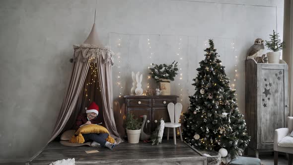 Boy with Glasses Sits in Festive Room with Christmas Tree Write Letter to Santa