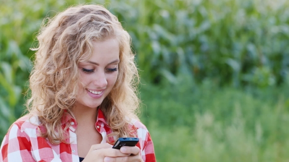 A Young Woman In The Countryside Using a Mobile Phone. It Stands On The Background Of Corn Fields