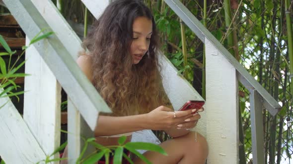 Young Woman with Long Curly Hair Types on Red Smartphone