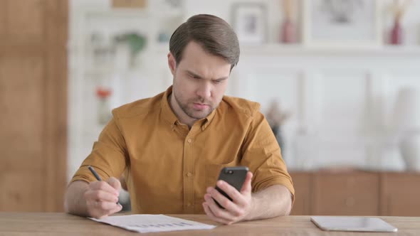 Young Man Looking at Phone while Writing on Paper in Office